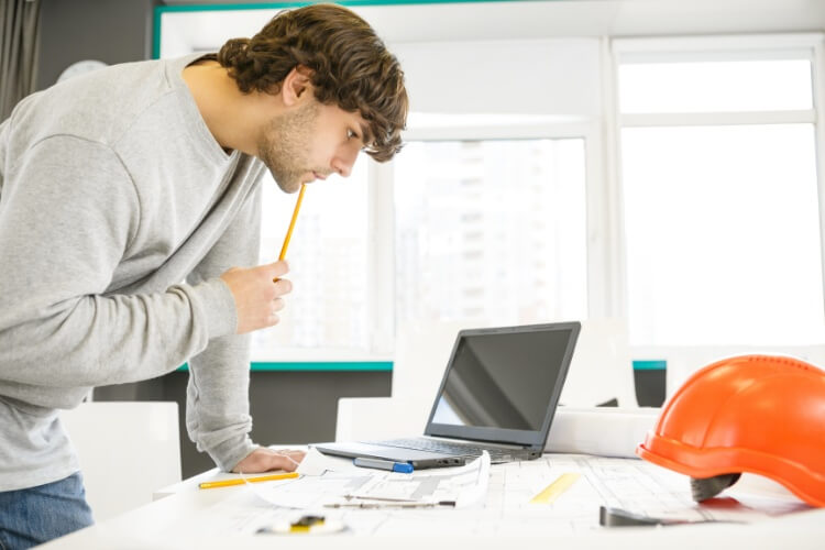 Artisan du bâtiment penché sur des plans avec un ordinateur et un casque de chantier, en train d'analyser des documents